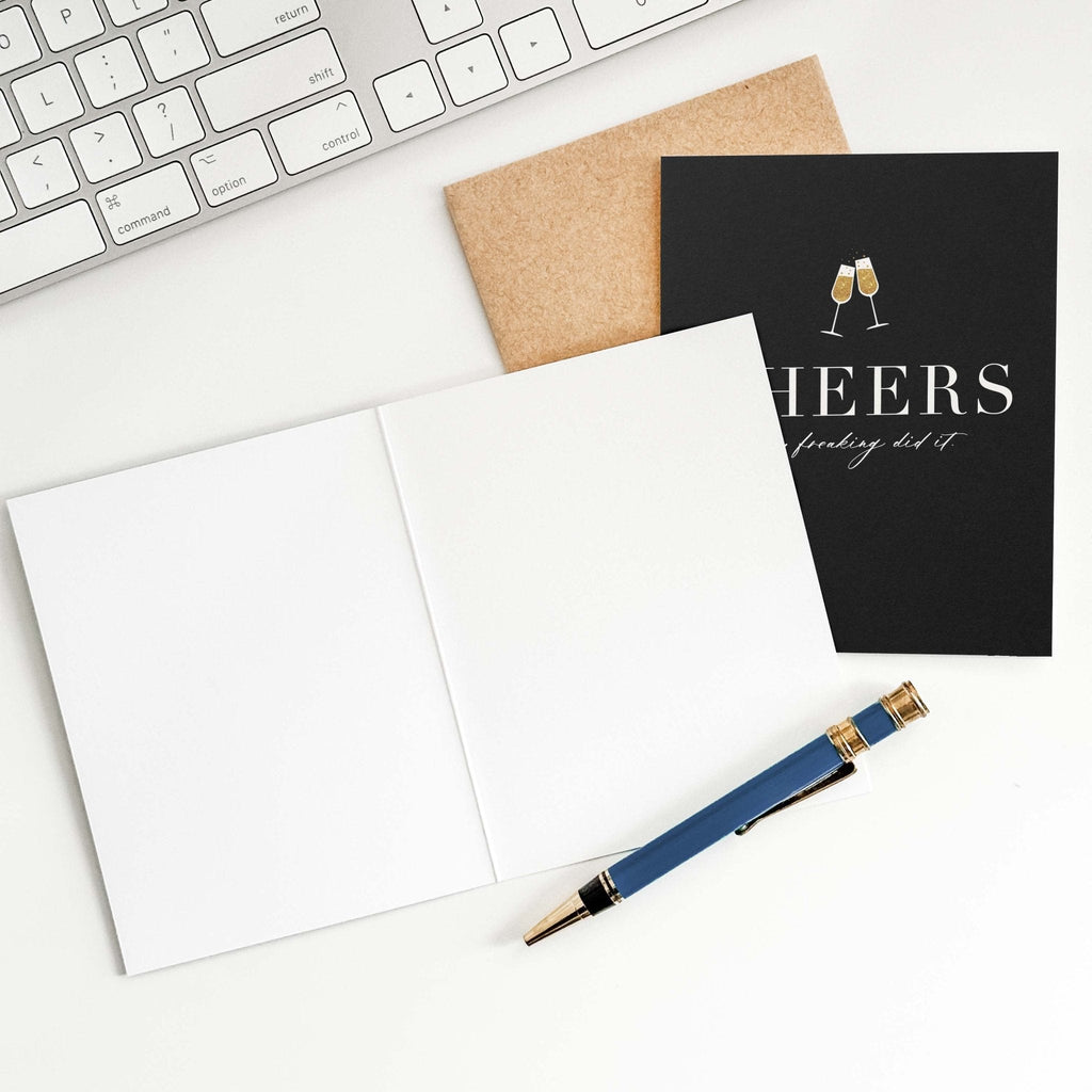 Cheers You Freaking Did It card by Blú Rose shown open with blank interior, kraft envelope and pen on a white desk