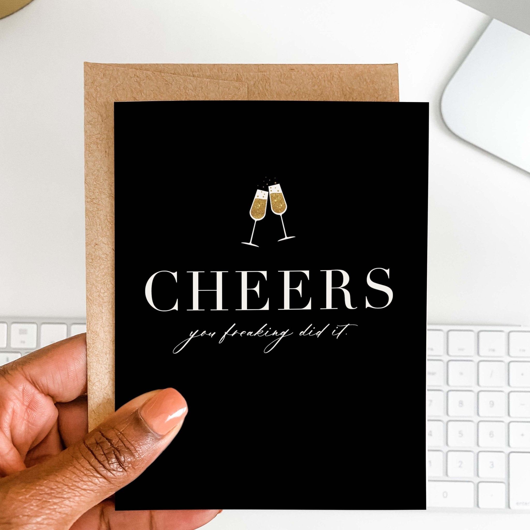 Cheers You Freaking Did It celebration card by Blú Rose held in hand over a white desk with keyboard and kraft envelope