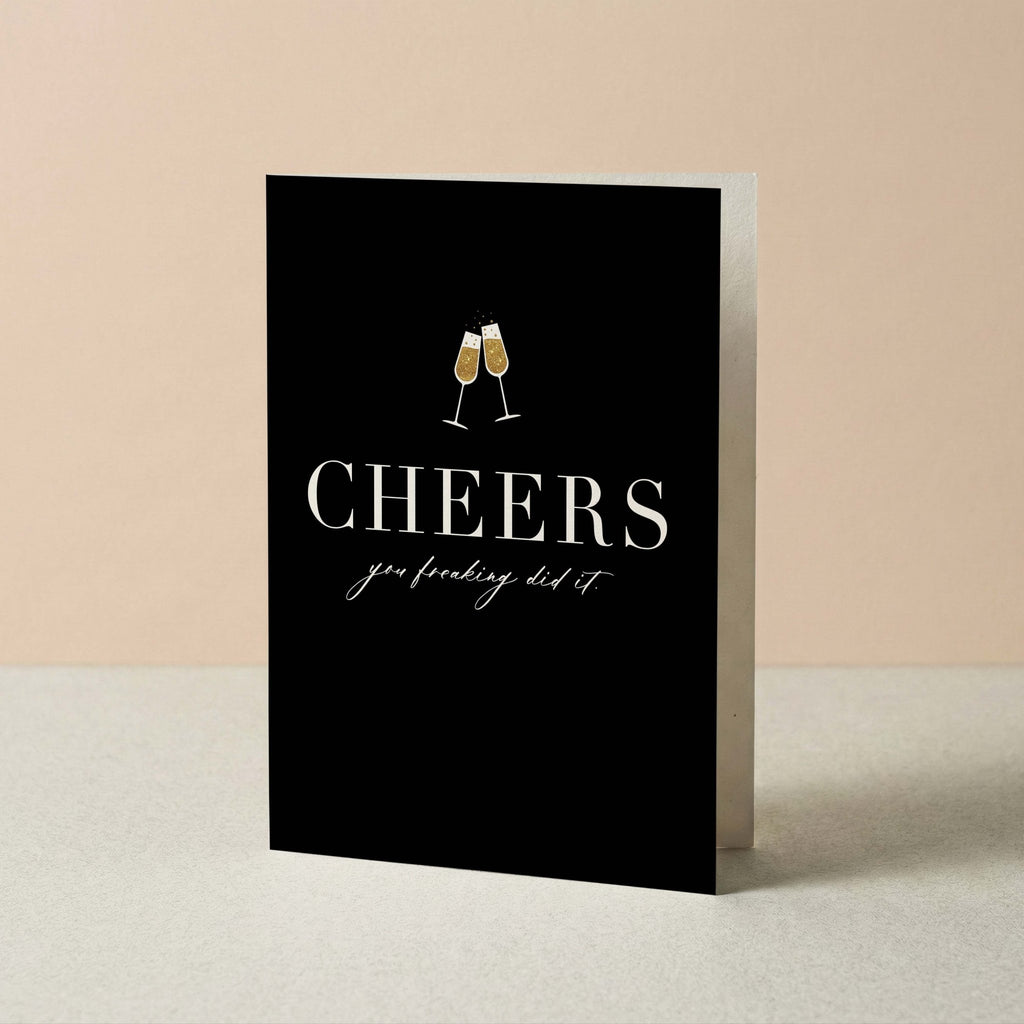 Cheers You Freaking Did It card by Blú Rose standing upright, black card with champagne cheers design on a neutral background