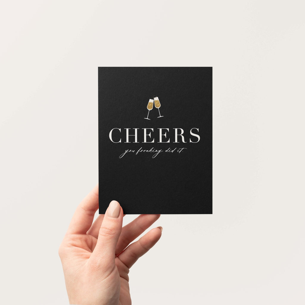 Cheers You Freaking Did It greeting card by Blú Rose held in hand against a neutral off-white background