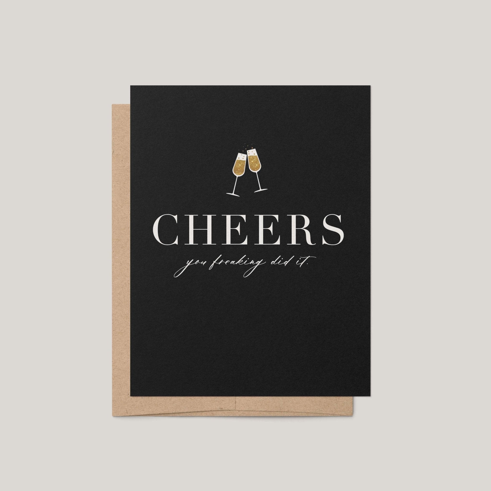 Cheers You Freaking Did It congratulations greeting card by Blú Rose, black card with gold champagne flutes and cheers text, shown with kraft envelope