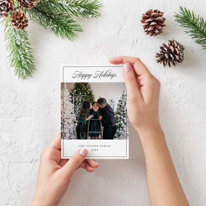 Hands holding modern family photo holiday card surrounded by pine greenery and seasonal accents.