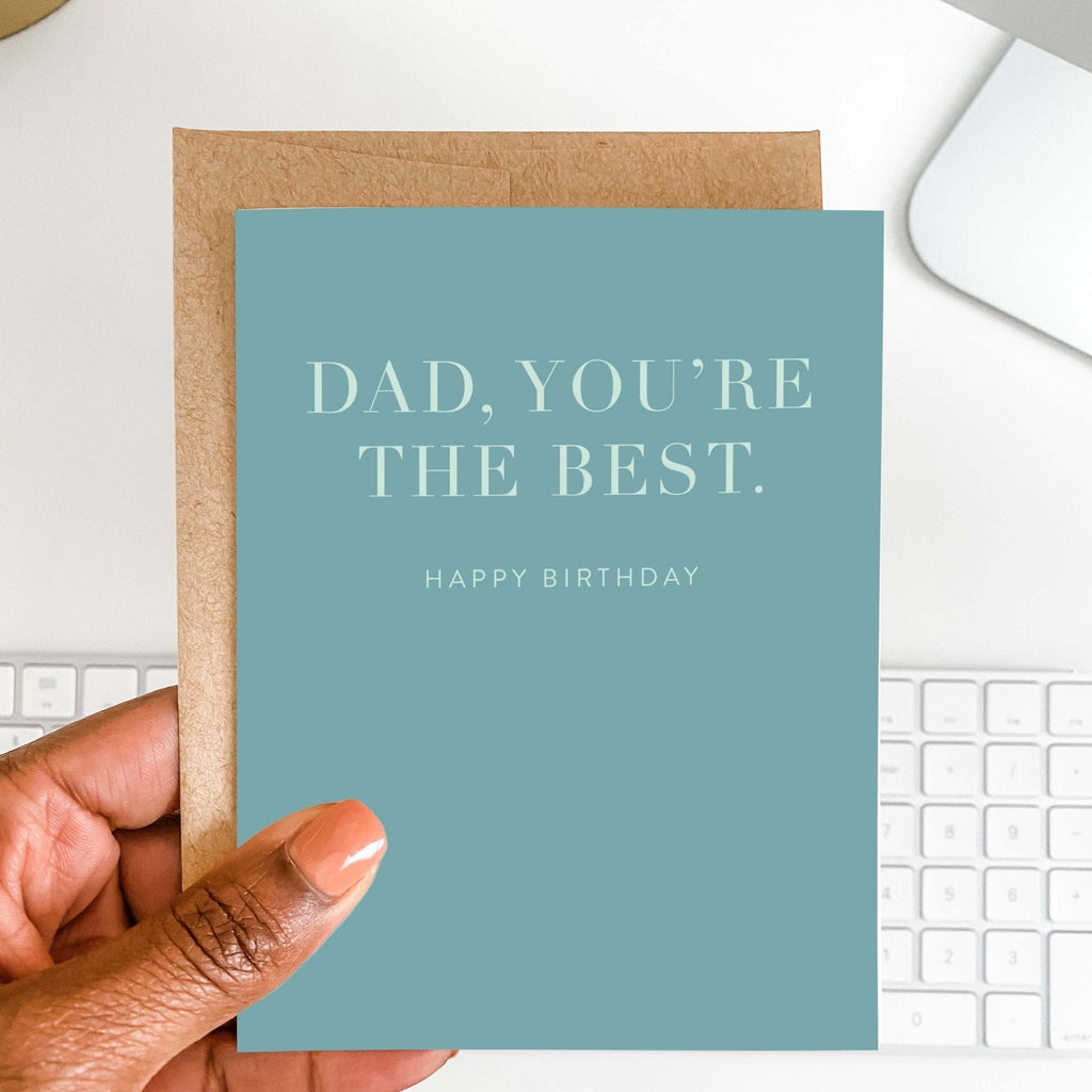 Dad You're the Best dad birthday card by Blú Rose held in hand over a white desk with keyboard and kraft envelope