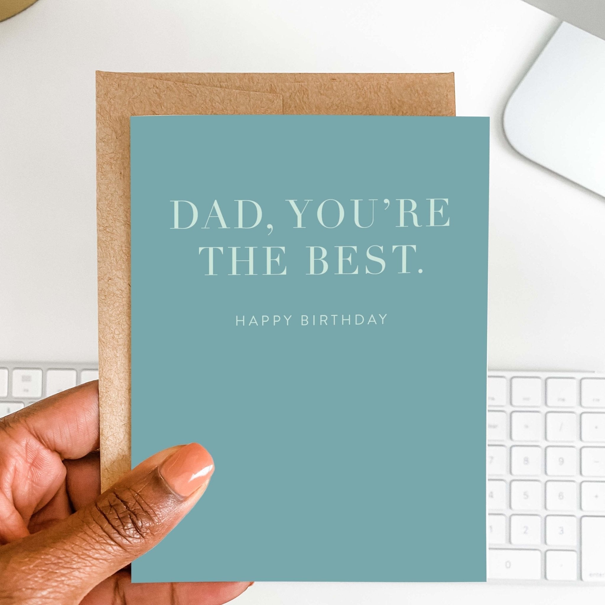 Dad You're the Best dad birthday card by Blú Rose held in hand over a white desk with keyboard and kraft envelope