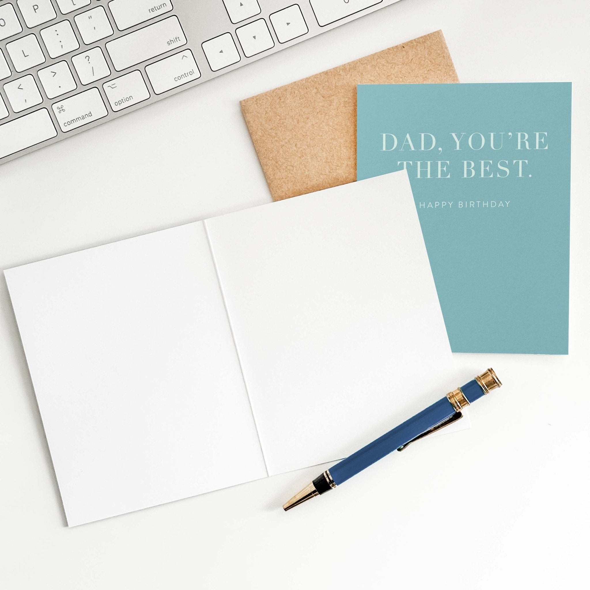 Dad You're the Best birthday card by Blú Rose shown open with blank interior, kraft envelope and pen on a white desk