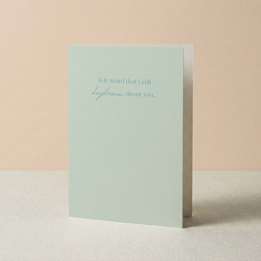 A pale mint green greeting card standing upright on a light surface against a soft peach background. The front of the card has a small printed phrase in muted blue-gray text reading, 'Is it weird that I still daydream about you.