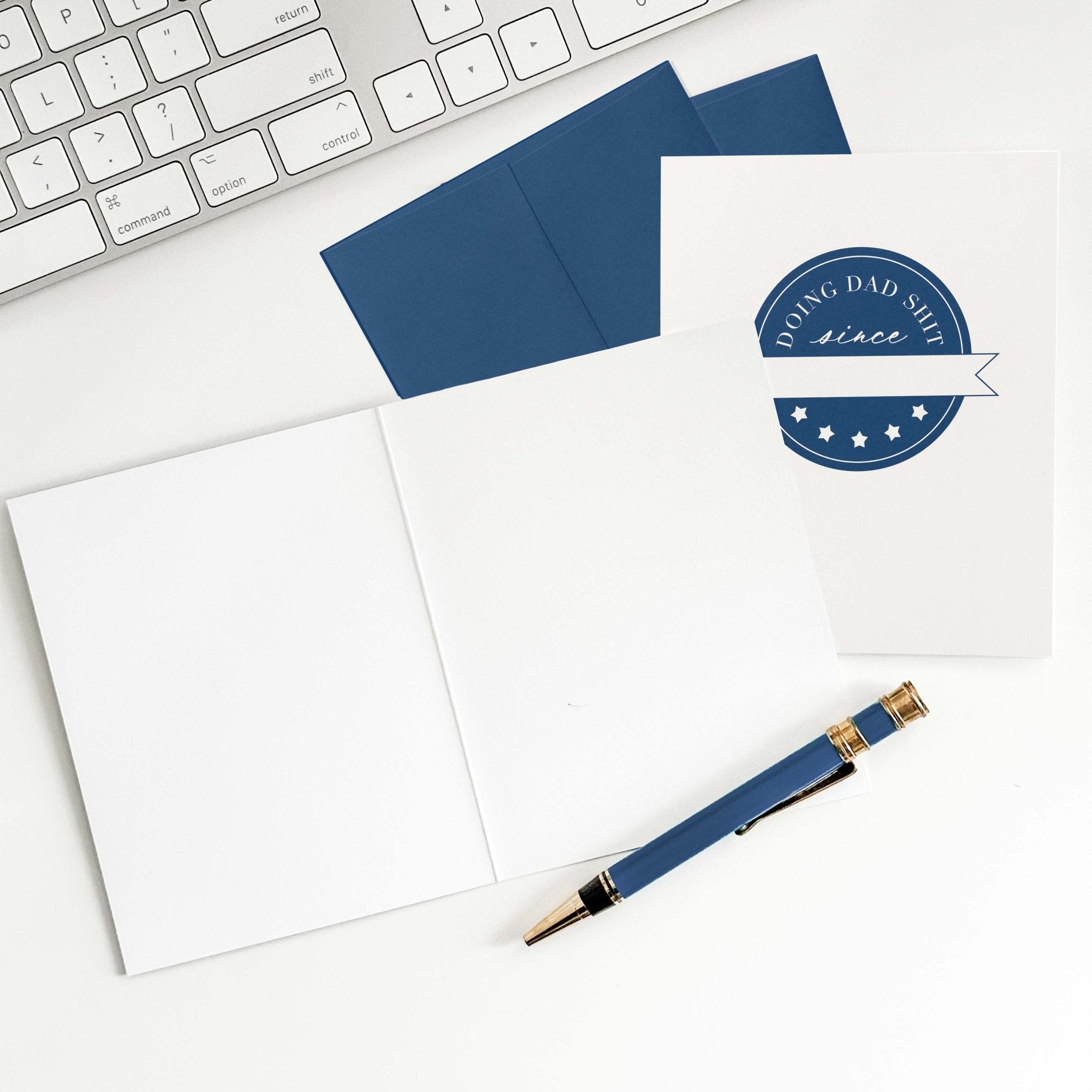 Doing Dad Shit card by Blú Rose shown open with blank interior, navy envelope and pen on a white desk