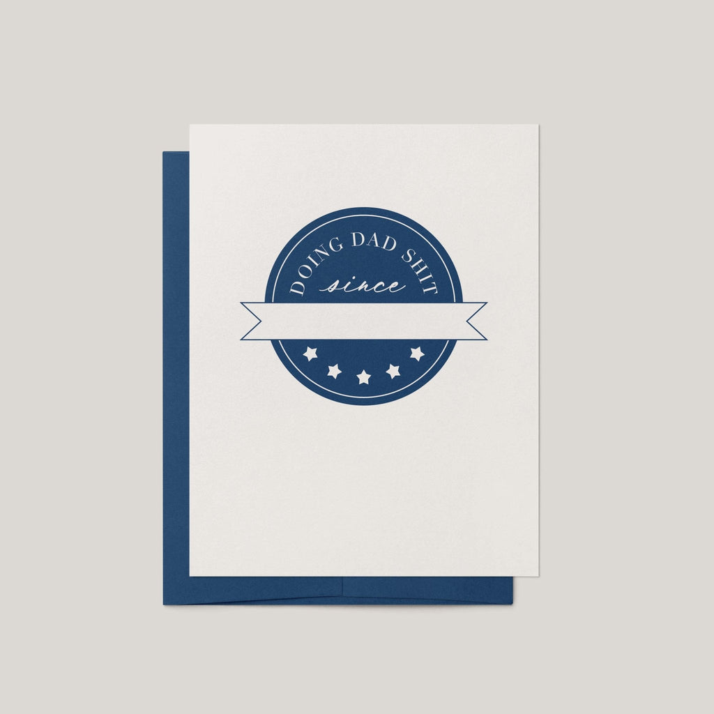 Doing Dad Shit funny Father's Day greeting card by Blú Rose, ivory card with navy badge crest design, shown with navy envelope