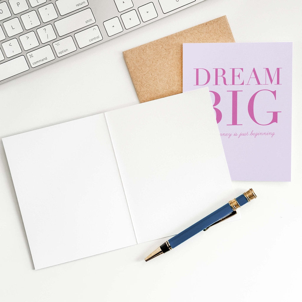Dream Big graduation card by Blú Rose shown open with blank interior, kraft envelope and pen on a white desk