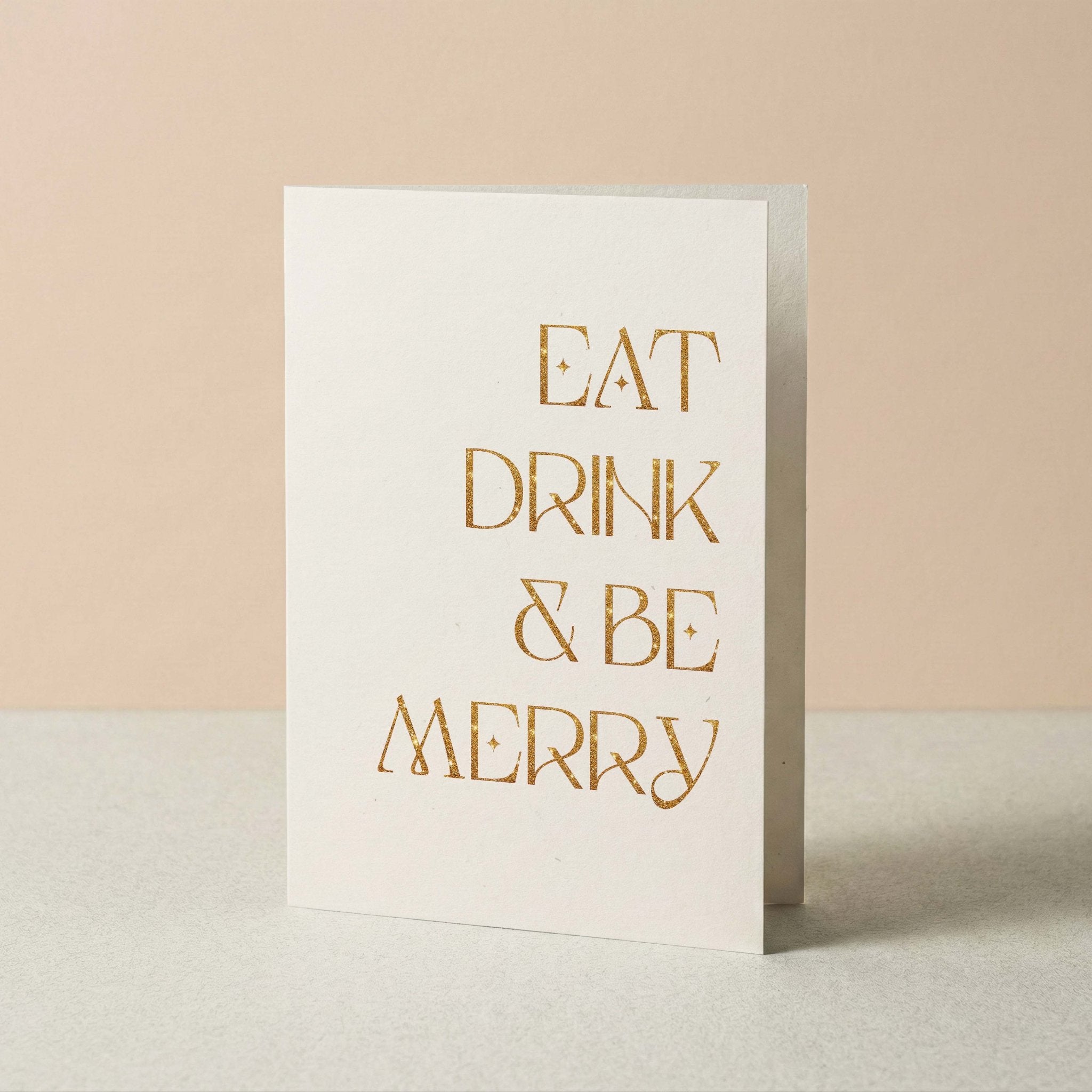 Eat, Drink & Be Merry Christmas greeting card by Blú Rose standing upright, ivory card with gold foil lettering on a neutral background
