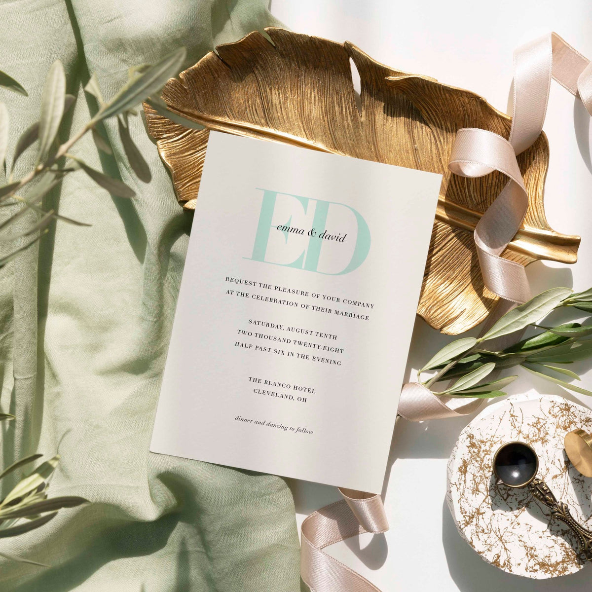 Emma wedding invitation by Blú Rose, cream card with a large mint 'ED' monogram, styled on a gold leaf dish with sage linen and ribbon