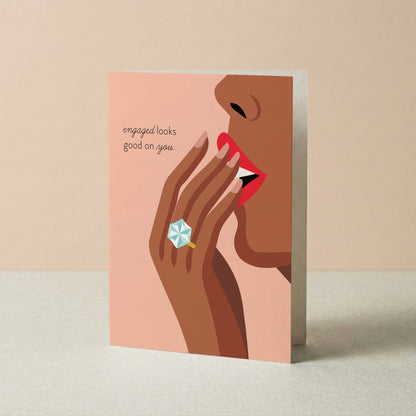 Greeting card with illustration of a hand with a ring and text 'engaged looks good on you' on a beige background