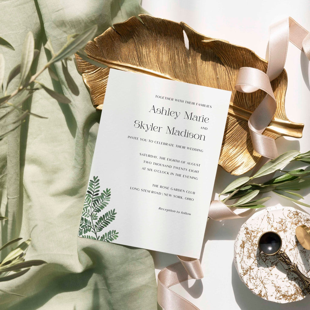 Fern wedding invitation by Blú Rose, white card with a green fern illustration and serif names, styled on a gold leaf dish with sage linen and ribbon