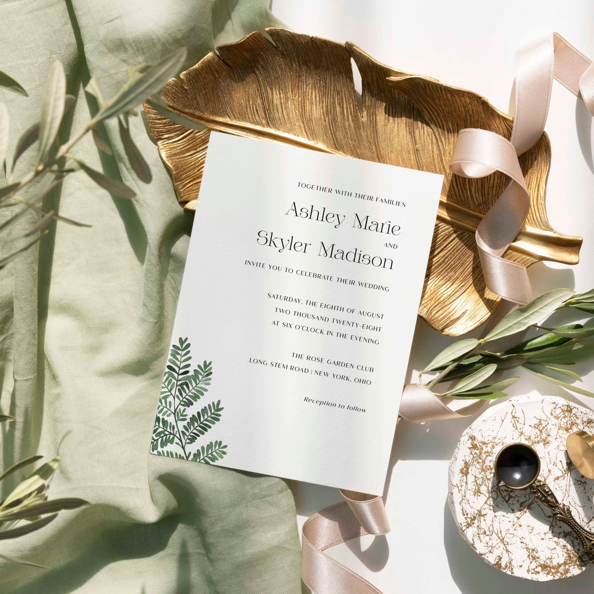 Fern wedding invitation by Blú Rose, white card with a green fern illustration and serif names, styled on a gold leaf dish with sage linen and ribbon