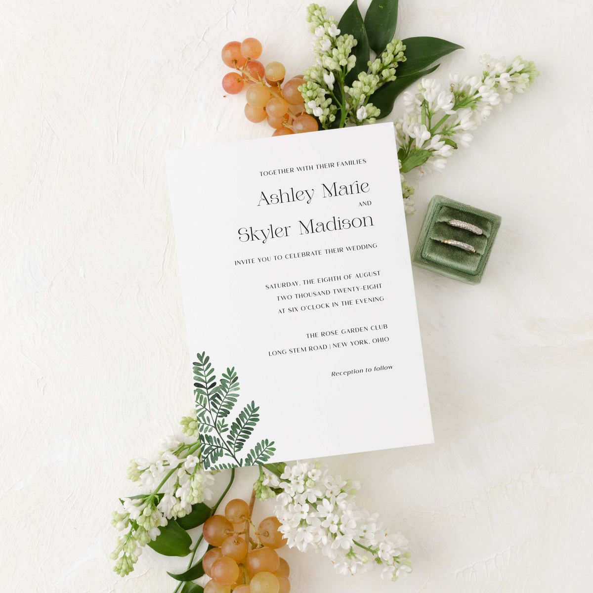 Fern wedding invitation by Blú Rose, white card with a green fern illustration and serif names, styled with white lilacs, grapes, and wedding rings