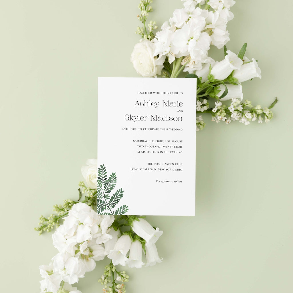 Fern wedding invitation by Blú Rose, white card with a green fern illustration and serif names, styled on a sage background with white blooms