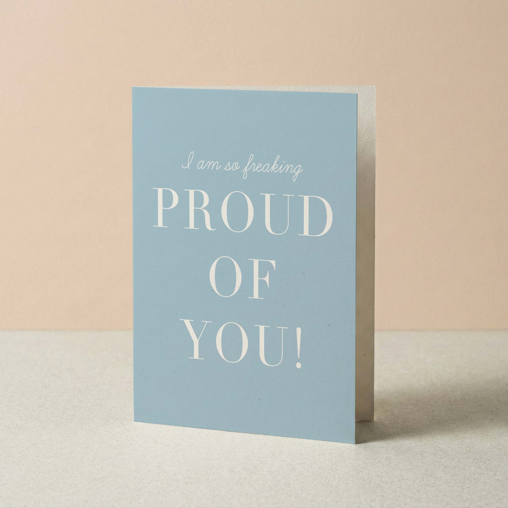 Freaking Proud of You card by Blú Rose standing upright, dusty blue card with white typography on a neutral background