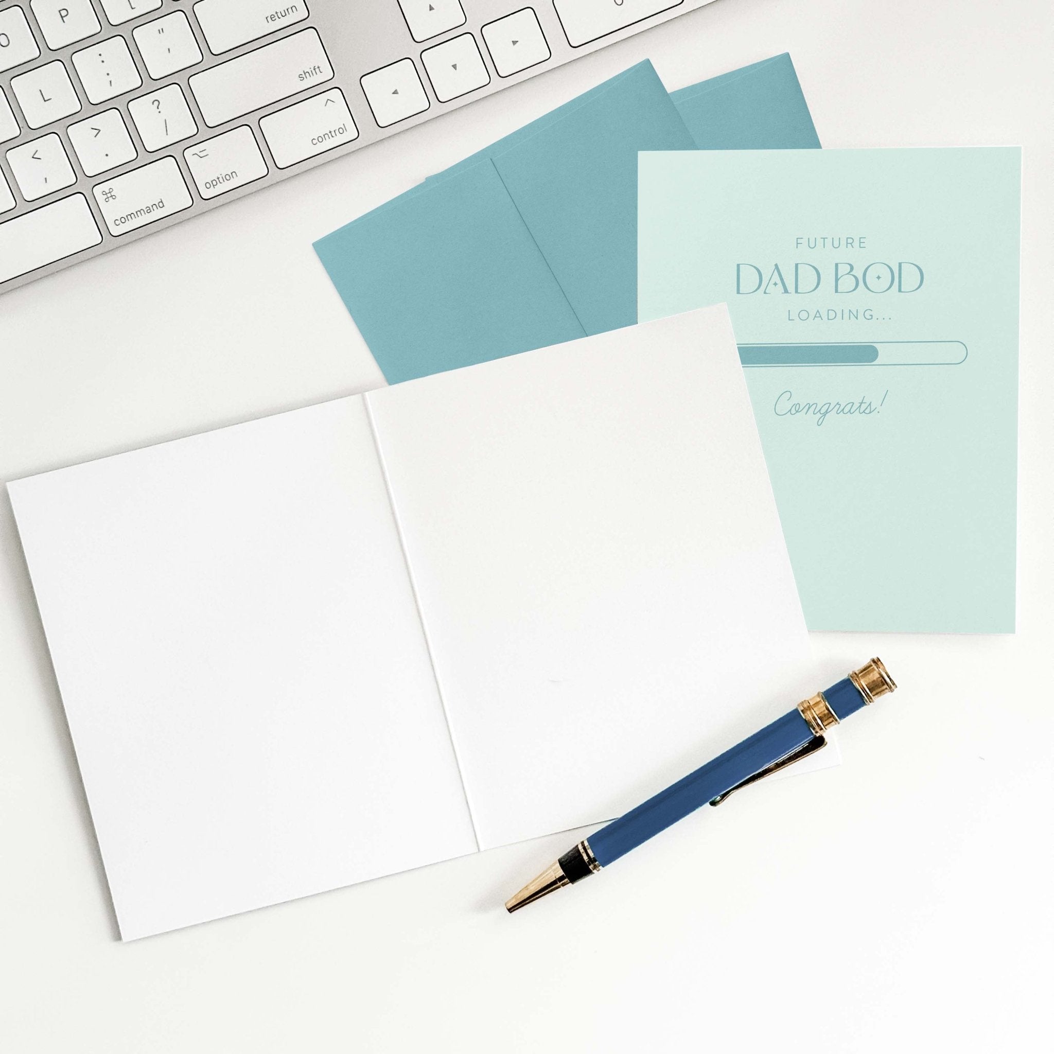 Future Dad Bod Loading card by Blú Rose shown open with blank interior, sage envelope and pen on a white desk