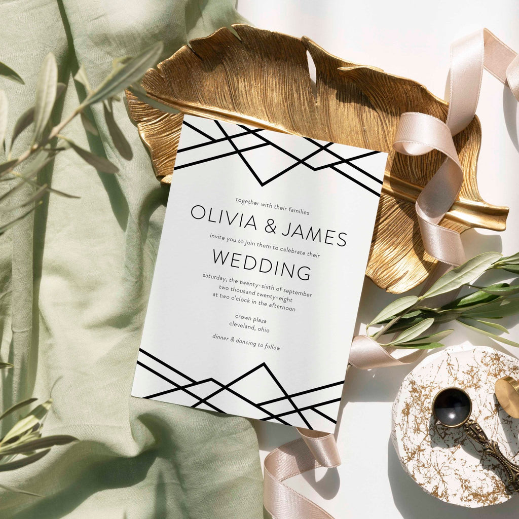 Geometric Art Deco wedding invitation by Blú Rose, white card with black geometric line-work, styled on a gold leaf dish with sage linen and ribbon