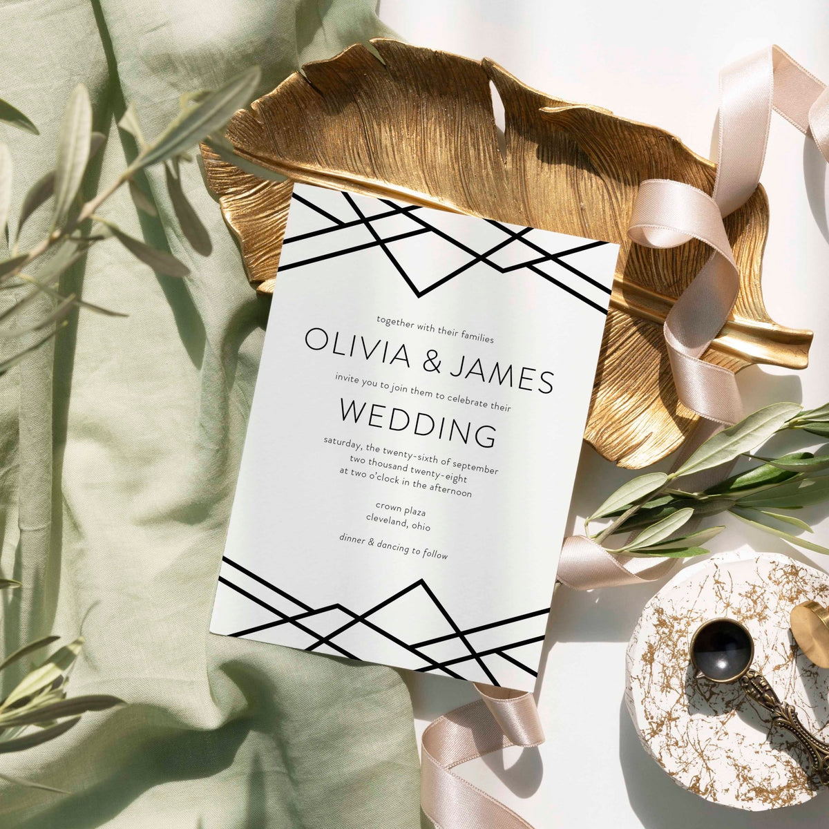 Geometric Art Deco wedding invitation by Blú Rose, white card with black geometric line-work, styled on a gold leaf dish with sage linen and ribbon