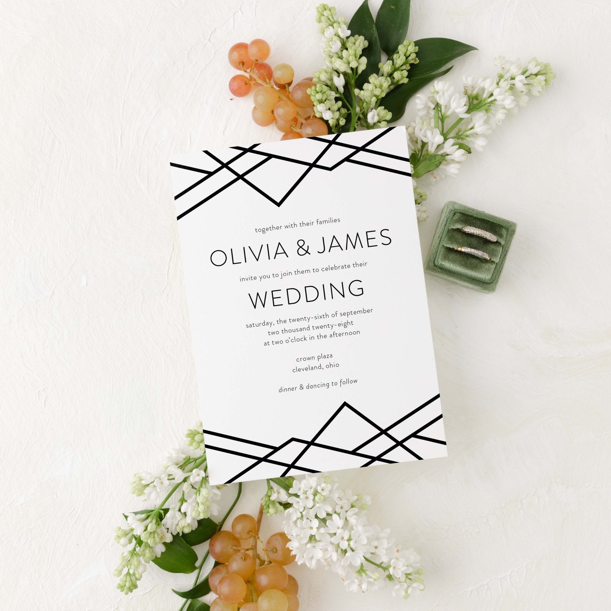 Geometric Art Deco wedding invitation by Blú Rose, white card with black geometric line-work and serif names, styled with white lilacs, grapes, and wedding rings