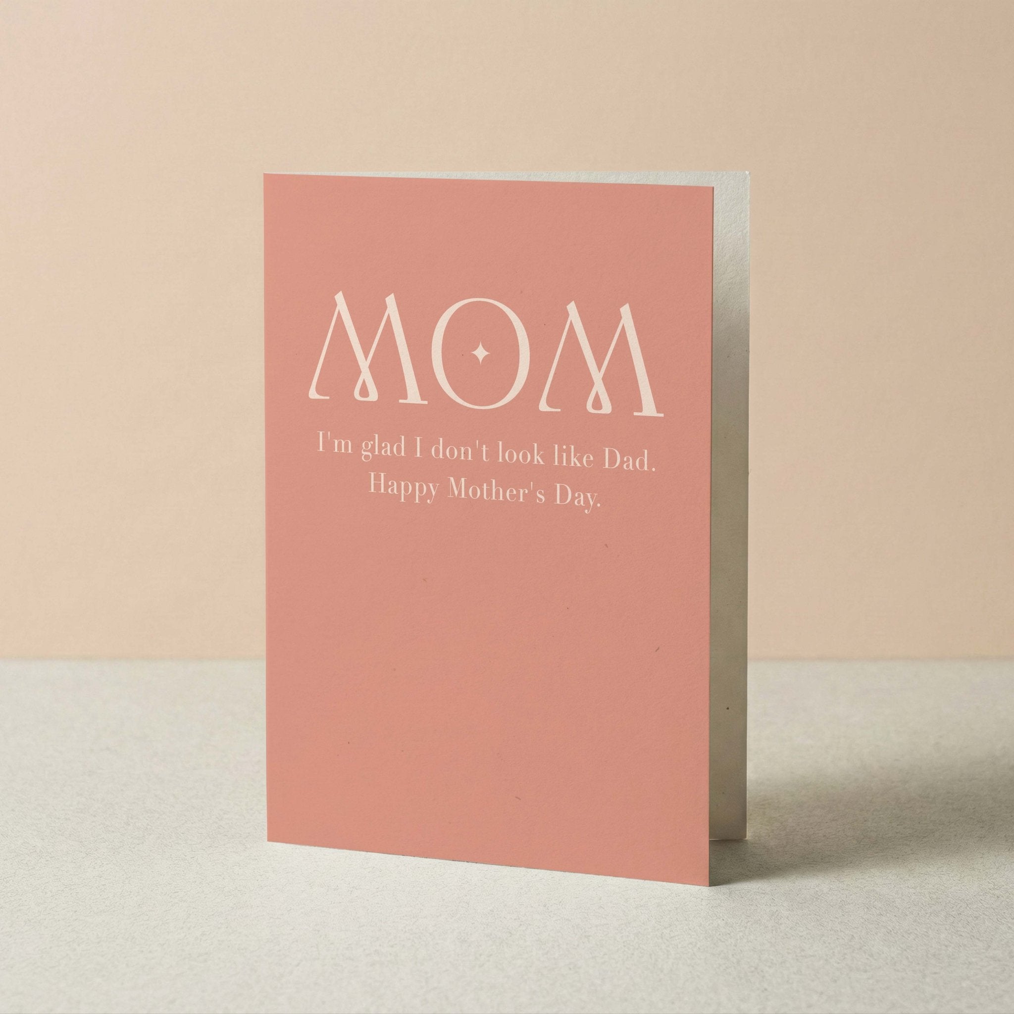 A pink greeting card standing upright on a light surface against a soft beige background. The card has white text reading “MOM” with smaller text below that says “I’m glad I don’t look like Dad.