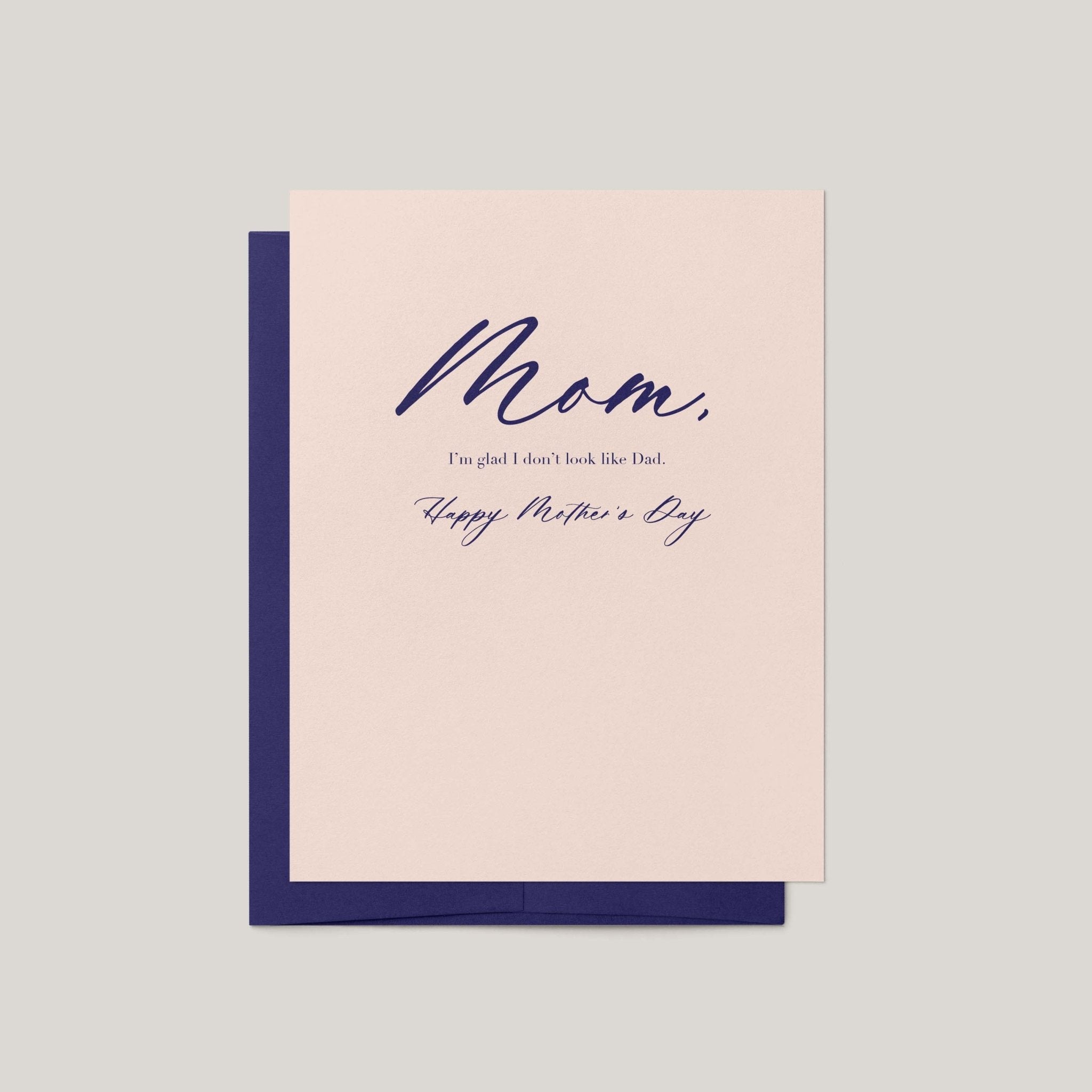 A Mother's Day greeting card with a pale blush pink front and navy blue envelope, featuring the handwritten-style text 'Mom, I'm glad I don't look like Dad. Happy Mother's Day.