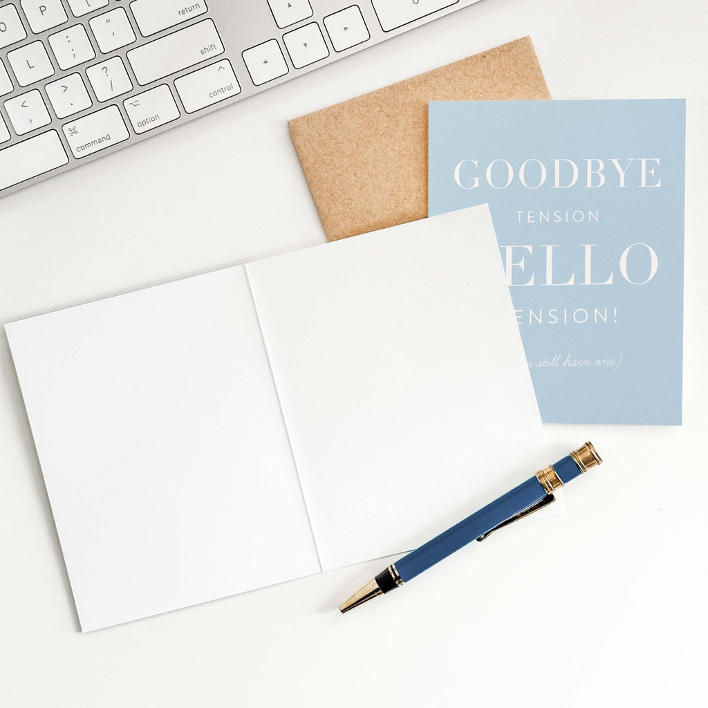 Goodbye Tension, Hello Pension retirement card by Blú Rose shown open with blank interior, kraft envelope and pen on a white desk