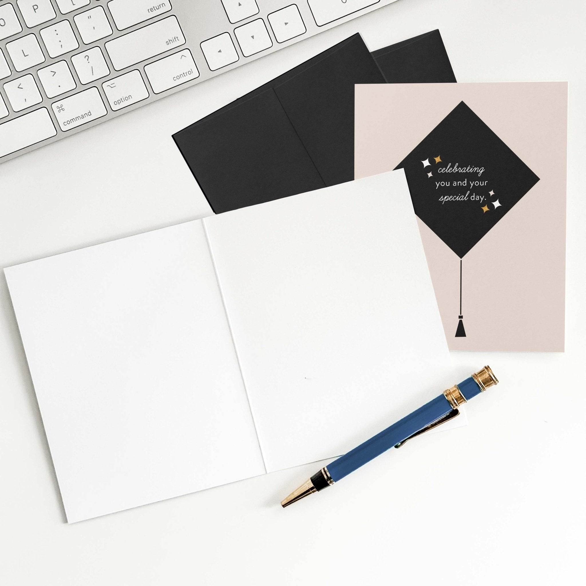 Grad Cap graduation card by Blú Rose shown open with blank interior, black envelope and pen on a white desk