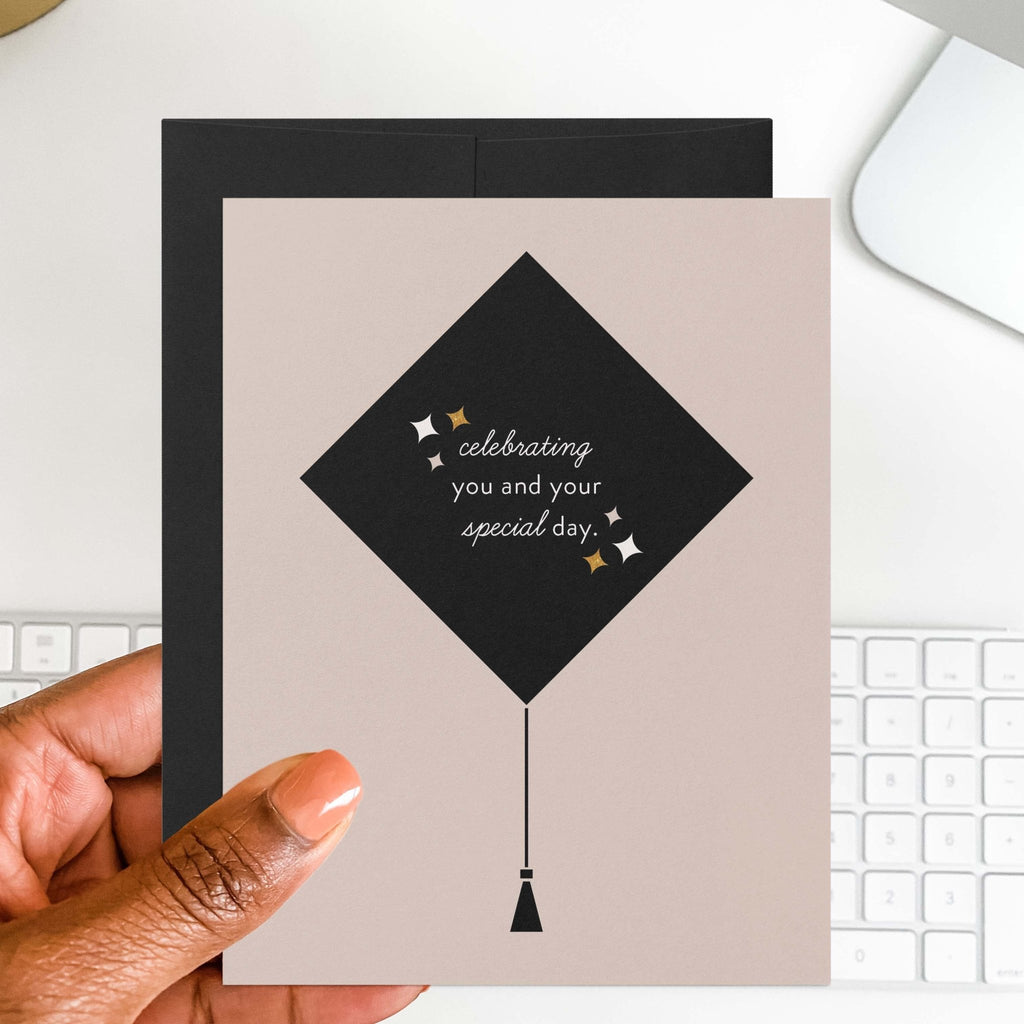 Grad Cap graduation card by Blú Rose held in hand over a white desk with keyboard and black envelope