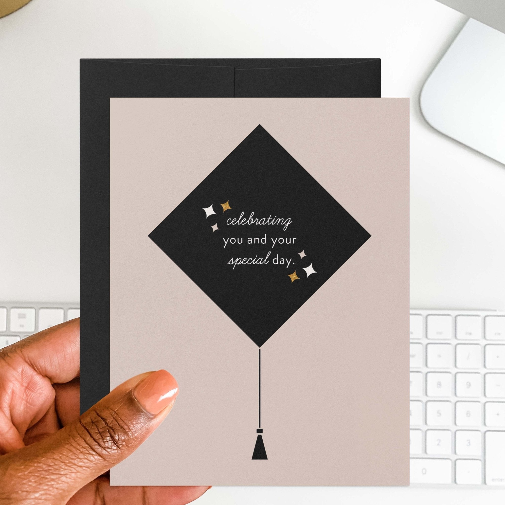 Grad Cap graduation card by Blú Rose held in hand over a white desk with keyboard and black envelope
