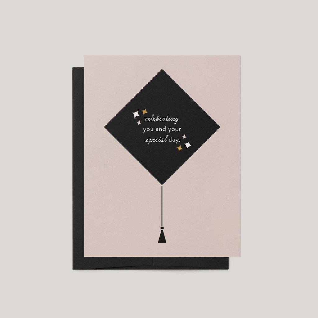 Grad Cap graduation greeting card by Blú Rose, blush card with black graduation cap illustration and celebrating you text, shown with black envelope