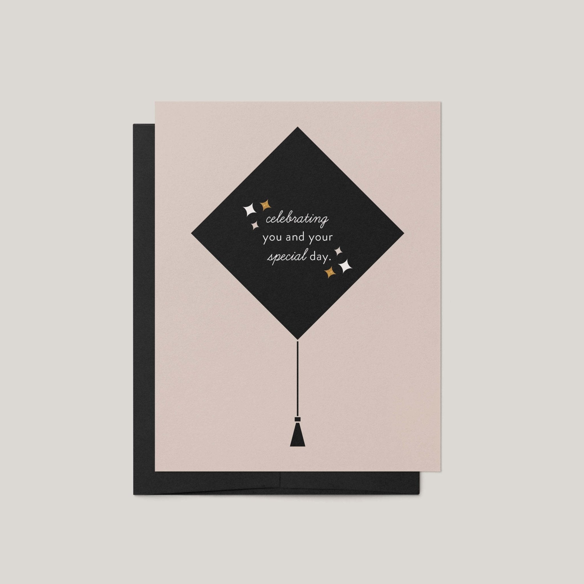 Grad Cap graduation greeting card by Blú Rose, blush card with black graduation cap illustration and celebrating you text, shown with black envelope