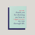 A greeting card with a light mint green front and a dark navy envelope behind it, featuring the printed message: 'mom, thank you for showing me how to improvise my way through life.' in navy and coral script.