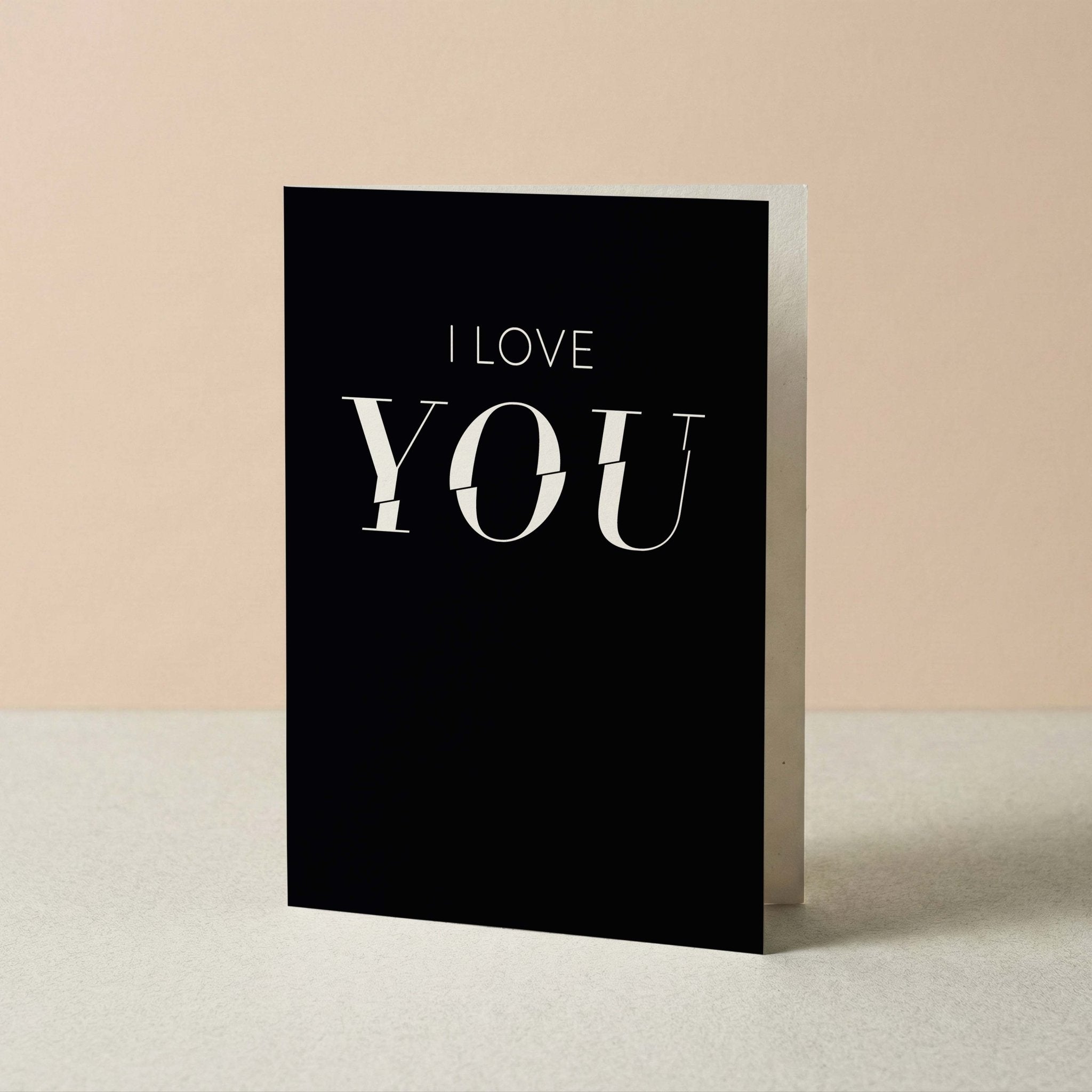 A black greeting card standing upright on a light surface against a warm beige background. The card features the text 'I LOVE YOU' in white, with 'YOU' in a large serif font and 'I LOVE' above it in a smaller sans-serif font.