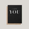A black greeting card with the words 'I LOVE YOU' printed in white, shown with a brown kraft envelope on a light gray background.