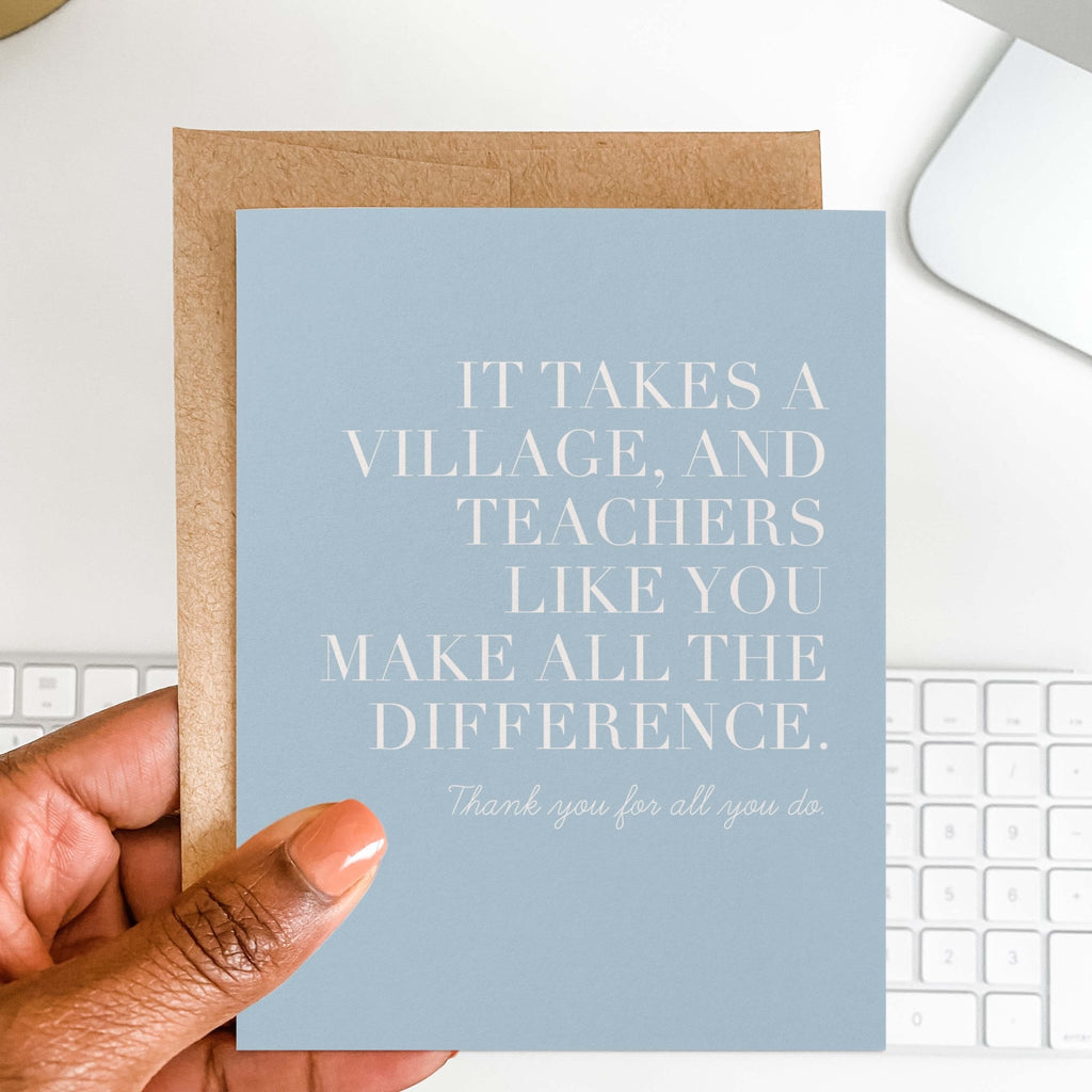 It Takes a Village teacher thank you card by Blú Rose held in hand over a white desk with keyboard and kraft envelope