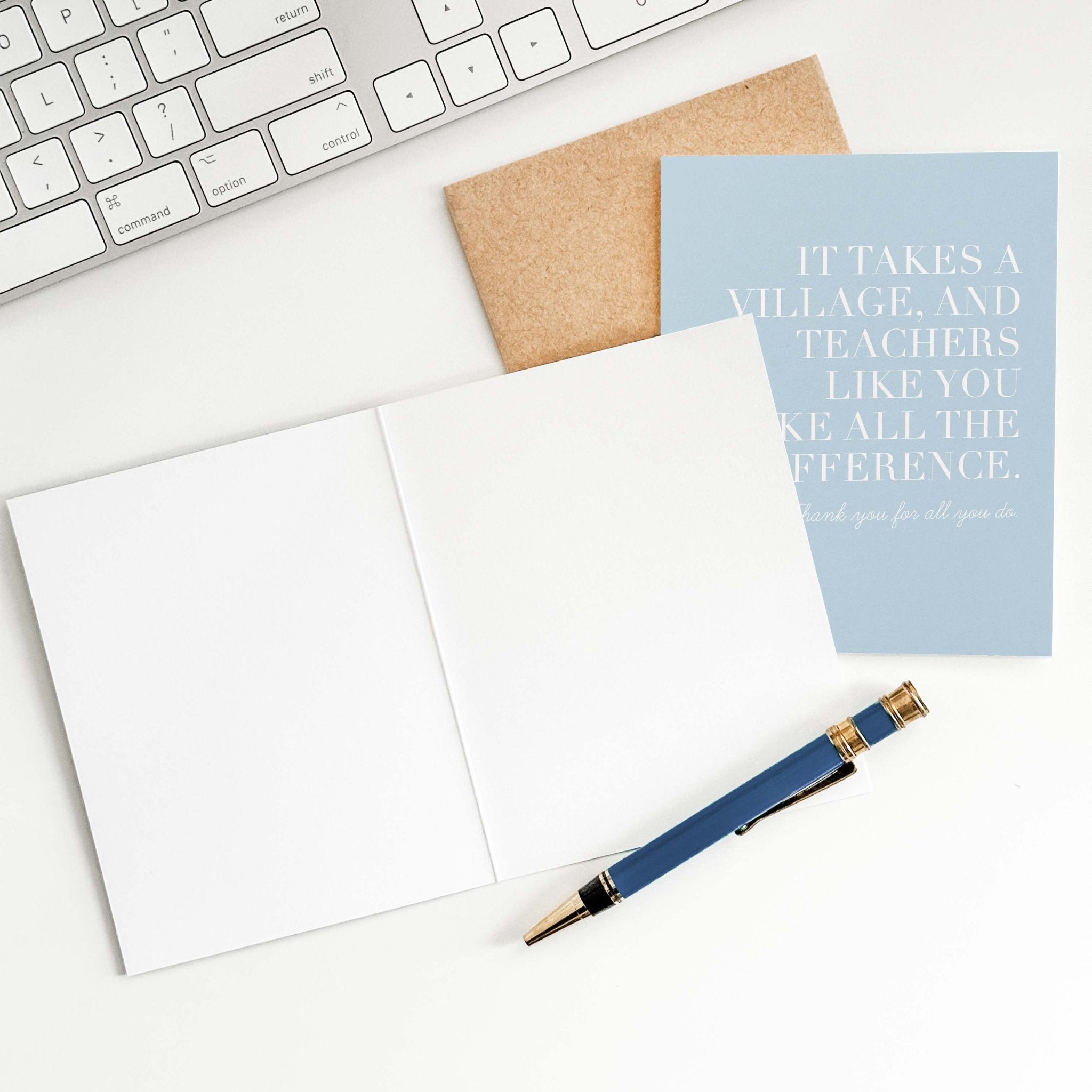 It Takes a Village teacher card by Blú Rose shown open with blank interior, kraft envelope and pen on a white desk