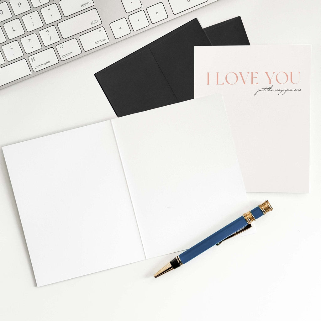 Just the Way You Are love card by Blú Rose shown open with blank interior, black envelope and pen on a white desk
