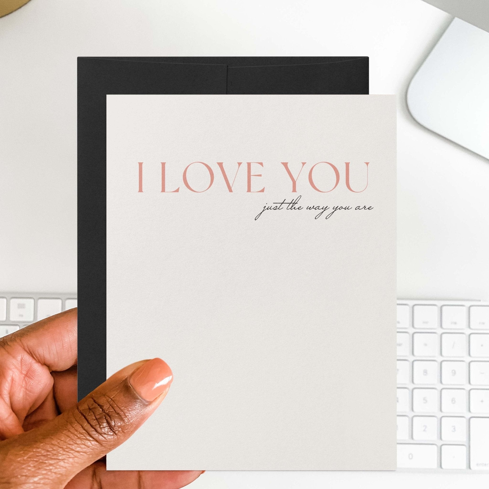 Just the Way You Are romantic card by Blú Rose held in hand over a white desk with keyboard and black envelope