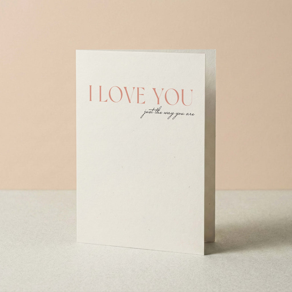 A minimalist white greeting card standing upright on a light surface against a soft peach background. The card reads 'I LOVE YOU' in a muted coral-pink serif font, with smaller script text below reading 'just the way you are'.