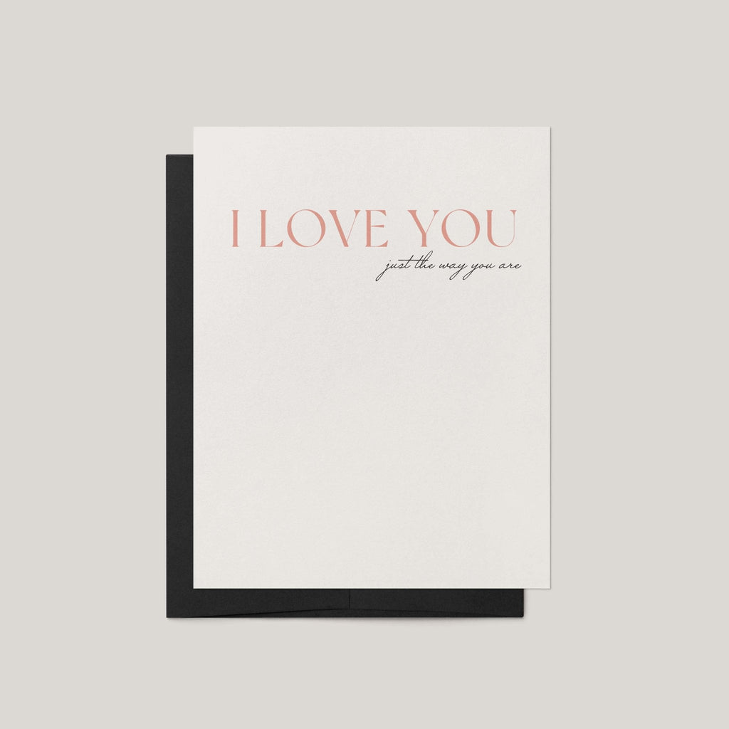 A minimalist greeting card with a white front and a black envelope, featuring the text 'I LOVE YOU just the way you are' in a soft coral and black script, set against a neutral light gray background.