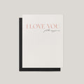 A minimalist greeting card with a white front and a black envelope, featuring the text 'I LOVE YOU just the way you are' in a soft coral and black script, set against a neutral light gray background.