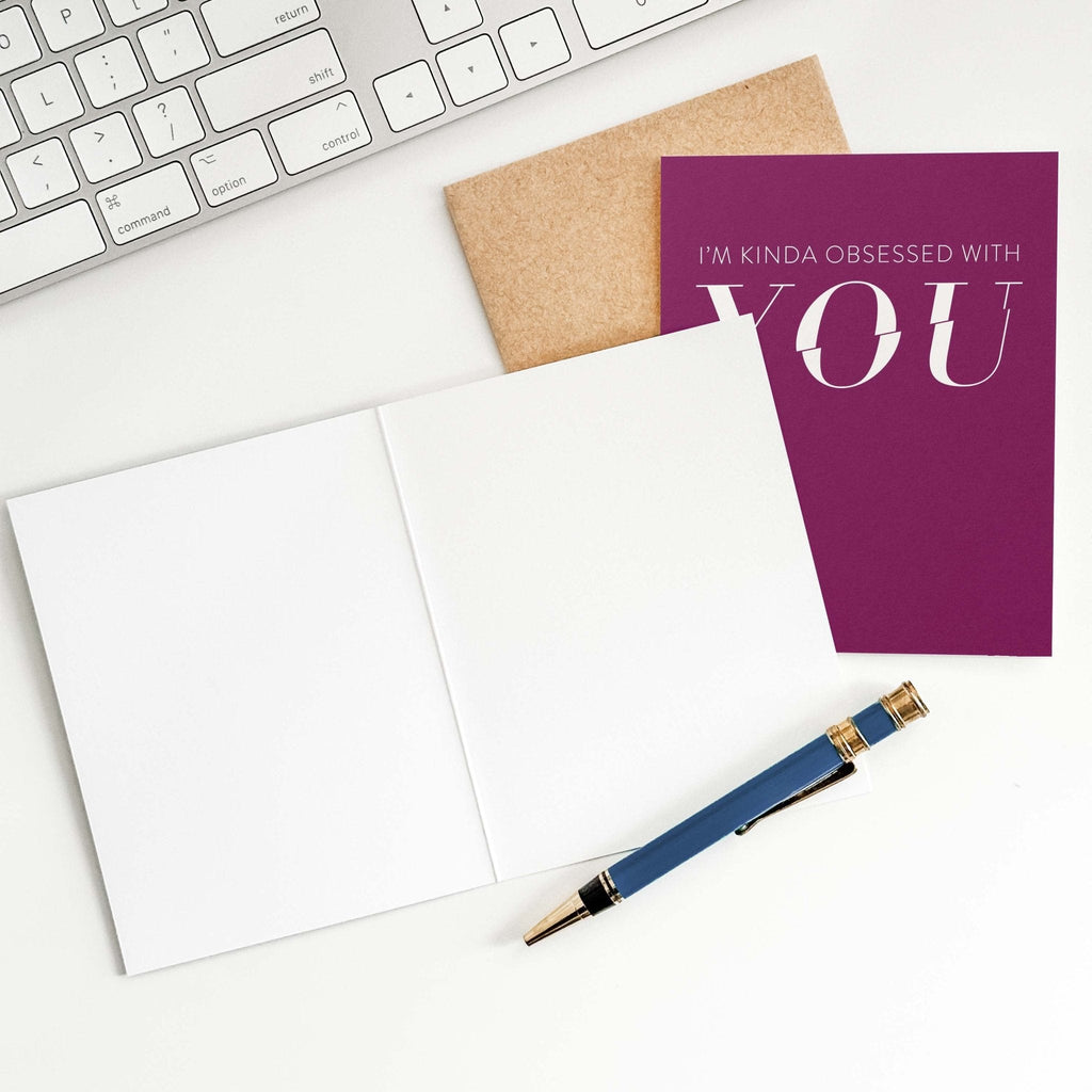 Kinda Obsessed love card by Blú Rose shown open with blank interior, kraft envelope and pen on a white desk