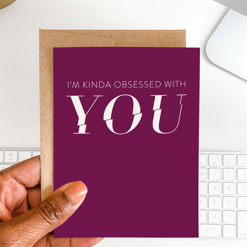 Kinda Obsessed romantic card by Blú Rose held in hand over a white desk with keyboard and kraft envelope