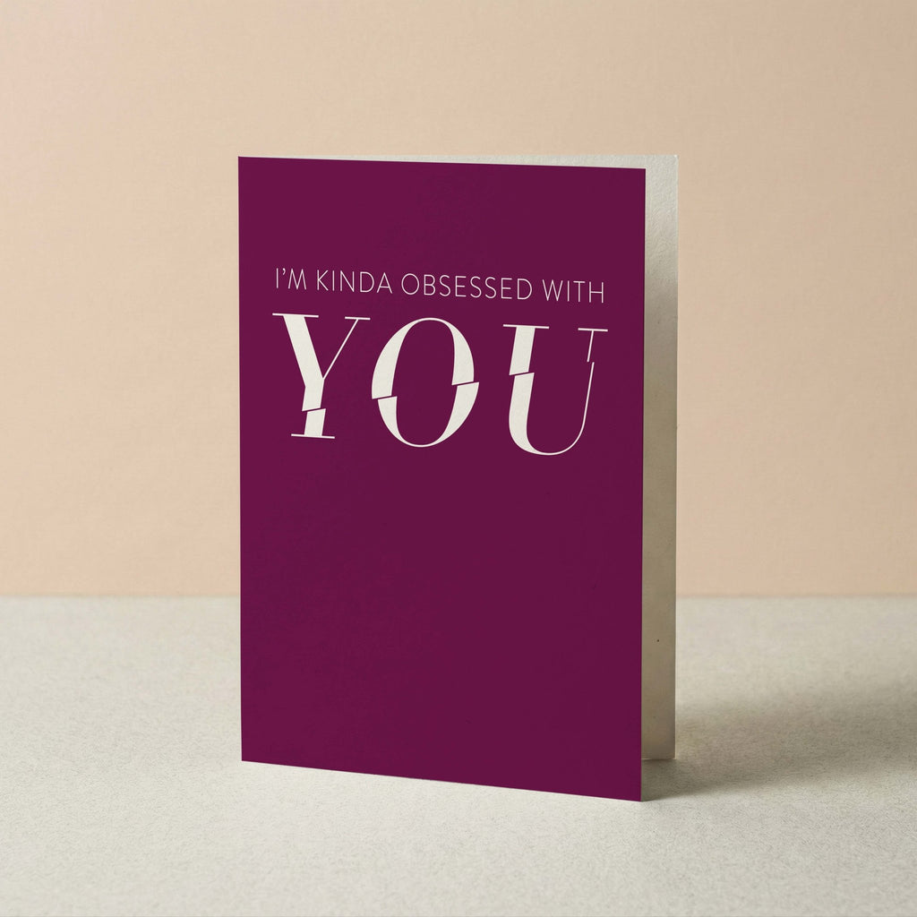 A purple greeting card with white text reading 'I'M KINDA OBSESSED WITH YOU' standing upright on a light surface against a beige background.