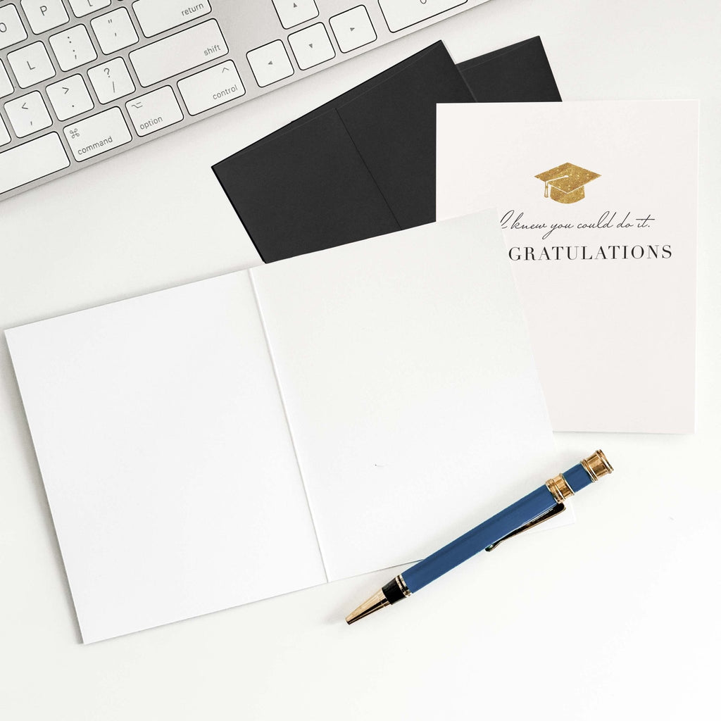 Knew You Could Do It graduation card by Blú Rose shown open with blank interior, black envelope and pen on a white desk