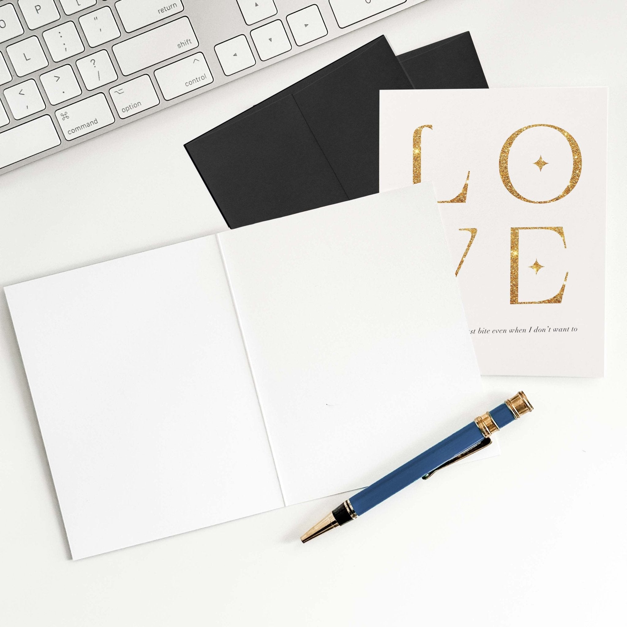 Love is Giving the Last Bite funny love card by Blú Rose shown open with blank interior, black envelope and pen on a white desk