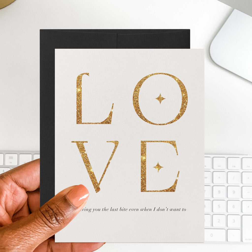 Love is Giving the Last Bite romantic card by Blú Rose held in hand over a white desk with keyboard and black envelope