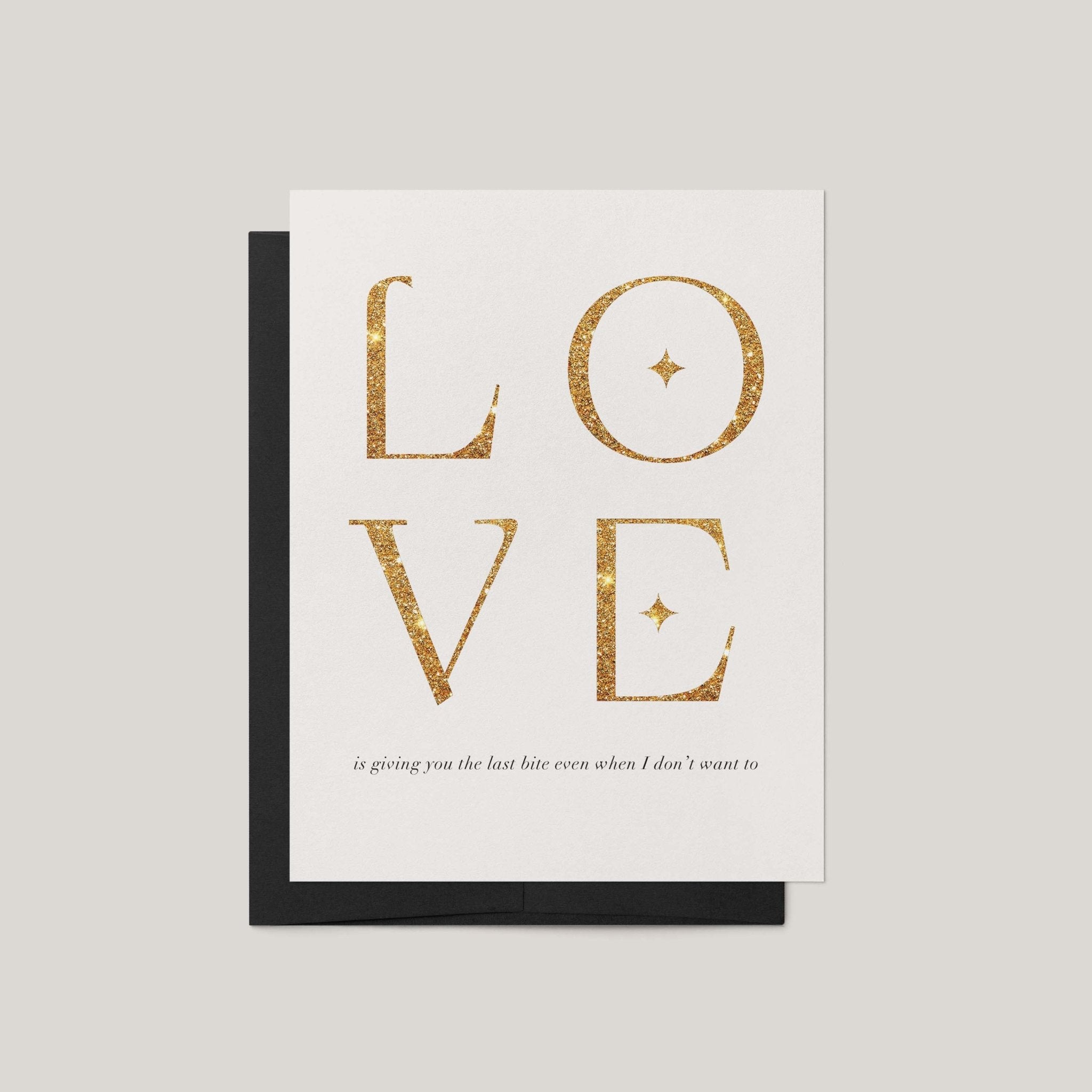 A greeting card with the word LOVE in large glittery gold letters on a white card, with a black envelope behind it, set against a light gray background.